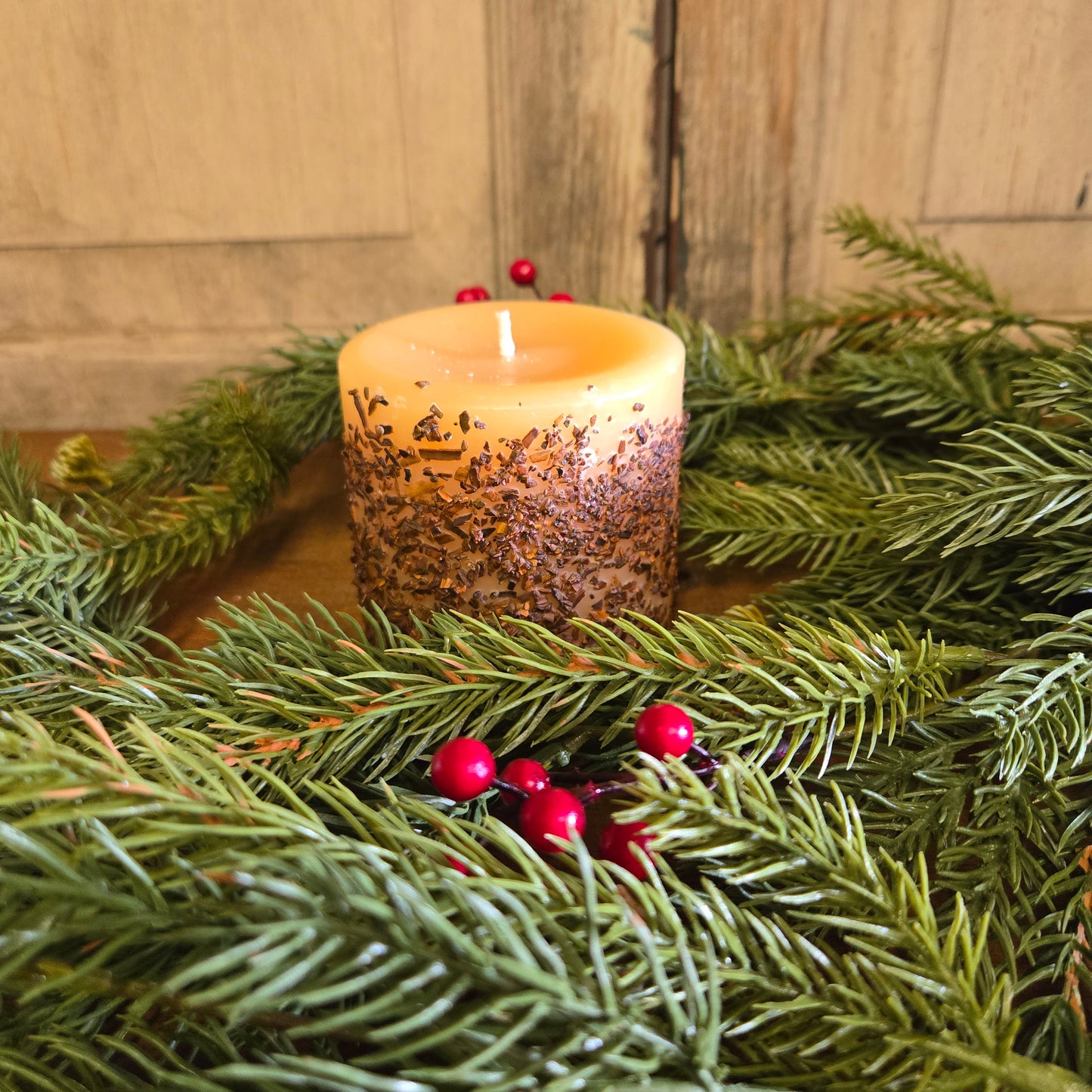 A snickerdoodle scented pillar candle with a vanilla and cinnamon scent, placed on a bed of artificial greenery with red berries.