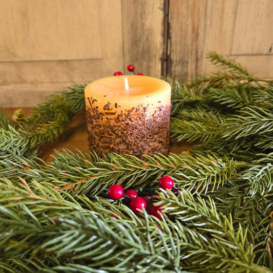 A snickerdoodle scented pillar candle with a vanilla and cinnamon scent, placed on a bed of artificial greenery with red berries.