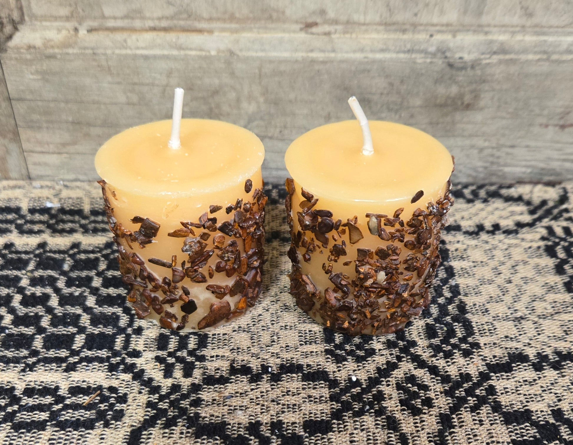 Two snickerdoodle scented votive candles with a warm glow, placed on a textured black mat, with a wooden background.