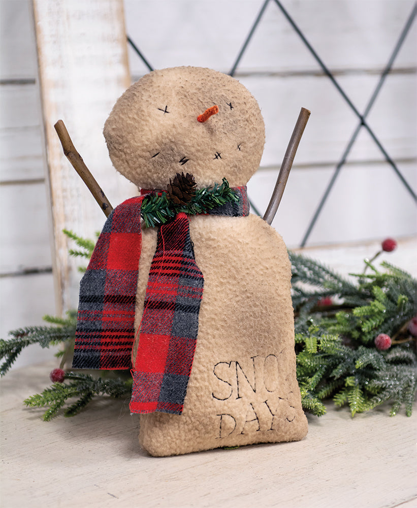 Stuffed snowman doll with a grungy fabric finish, orange carrot nose, plaid scarf, pine spray, and stick arms. It has 'Snow Days' text embroidered on its chest.
