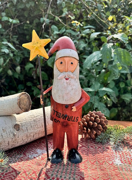 A decorative figurine of Santa Claus wearing a red suit and holding a yellow star, placed on a red surface with a greenery background.