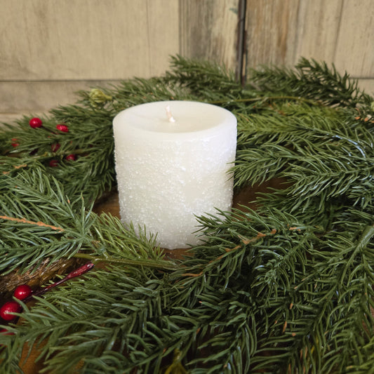 A white pillar candle surrounded by green pine branches and red berries, placed on a wooden surface.