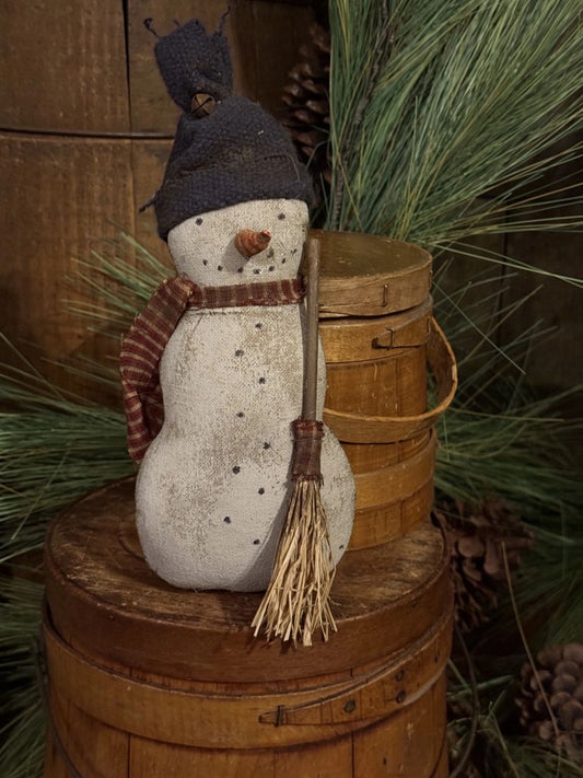 A rustic handmade snowman decoration wearing a dark knit hat and striped scarf, standing on a wooden barrel with pine branches and a small straw broom in a holiday setting.