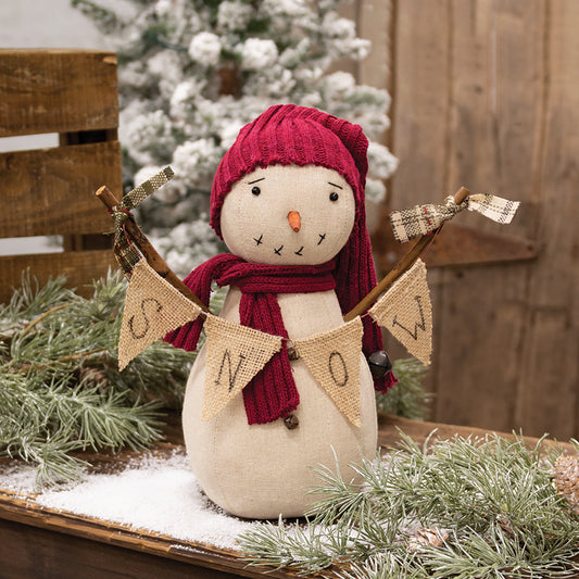 Stuffed snowman doll with a red hat and scarf, holding a burlap banner that reads 'snow', standing on a shelf with a Christmas tree and decorations in the background.
