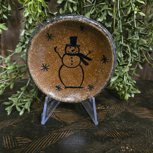 Decorative pottery plate with a snowman design on a wooden surface with greenery