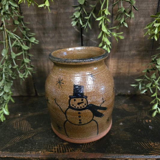 Stoneware crock with a snowman design on a wooden surface with green leaves.