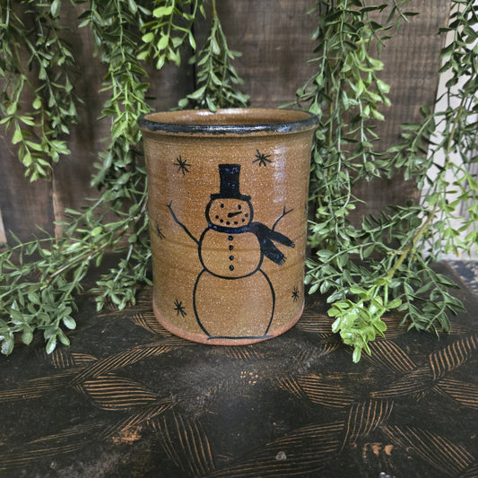 Cylindrical ceramic container with a snowman design on a wooden surface with greenery.