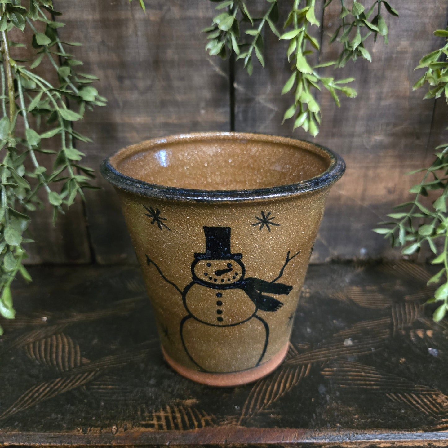 Brown ceramic tumbler with a snowman design on a wooden surface with greenery