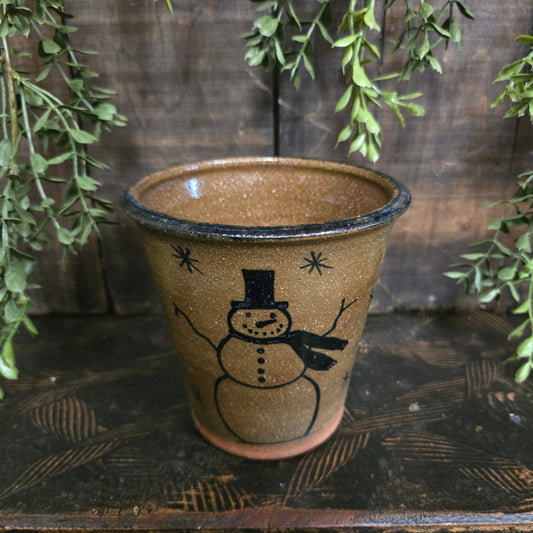 Brown ceramic tumbler with a snowman design on a wooden surface with greenery