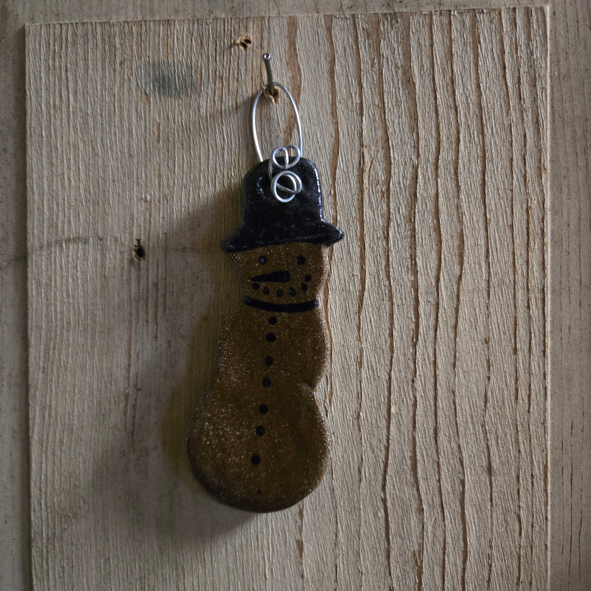 A rustic handcrafted snowman ornament hanging from a metal hook on a light wood plank background. The ornament features a brown glittery body with black painted facial features and a black hat, topped with a silver-colored wire hanger.