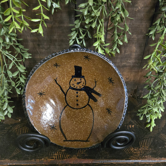 Decorative plate with a snowman on a brown background, surrounded by green leaves.