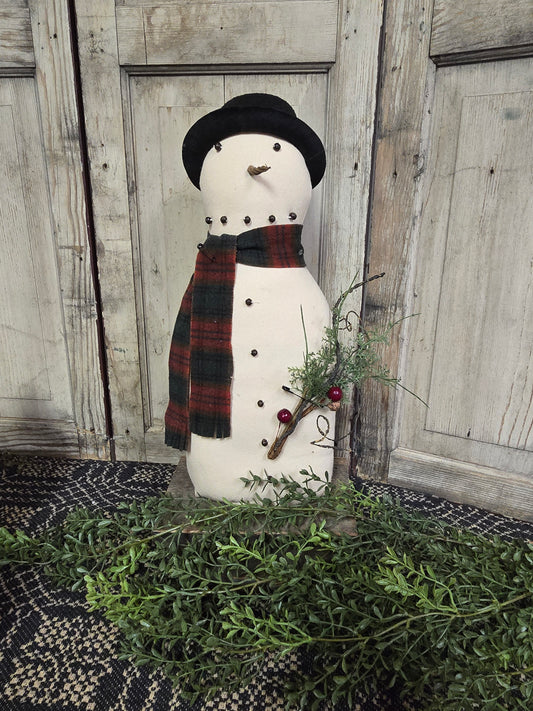 Snowman figurine with a black hat, plaid scarf, and small wreath against a wooden wall.