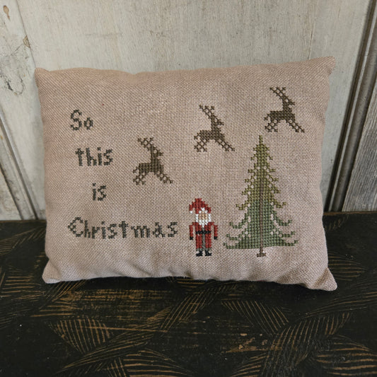 Decorative pillow with cross-stitched design featuring reindeer and 'So this is Christmas' text, placed against a textured wall.