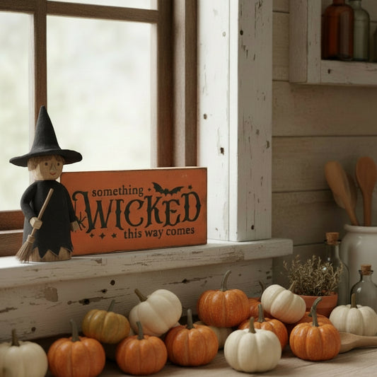 Decorative setup with pumpkins, a witch figurine, and a sign reading 'Something Wicked This Way Comes' on a rustic shelf.