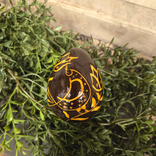 A brown ceramic egg with red and yellow sgraffito patterns, placed on a green plant, indicative of a handcrafted piece in a home decor setting.