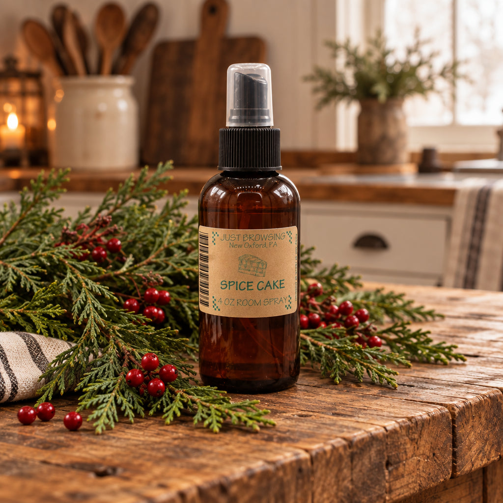 An amber glass room spray bottle labeled 'Spice Cake' stands on a rustic wooden table surrounded by evergreen branches and red berries in a warm kitchen setting.