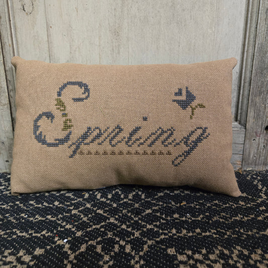 A rectangular cross stitch pillow with a floral design and the word 'Spring' embroidered on it, displayed on a textured surface.