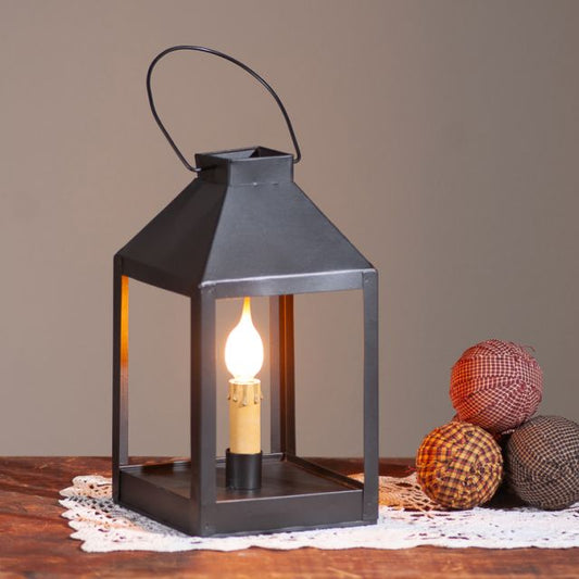A smokey black metal square table accent light with a warm glow, displayed on a wooden surface with decorative items around it.