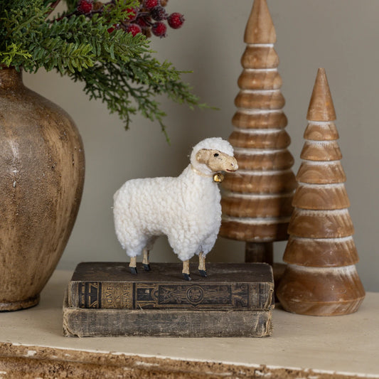 A small white woolly sheep figurine with a gold bell stands on top of two vintage books on a shelf, styled with a large vase of greenery and two wooden Christmas trees in the background.