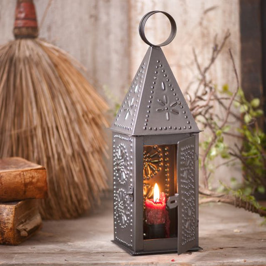 A black square-shaped metal lantern with geometric patterns, displaying a lit candle inside, placed on a wooden surface with a rustic background.
