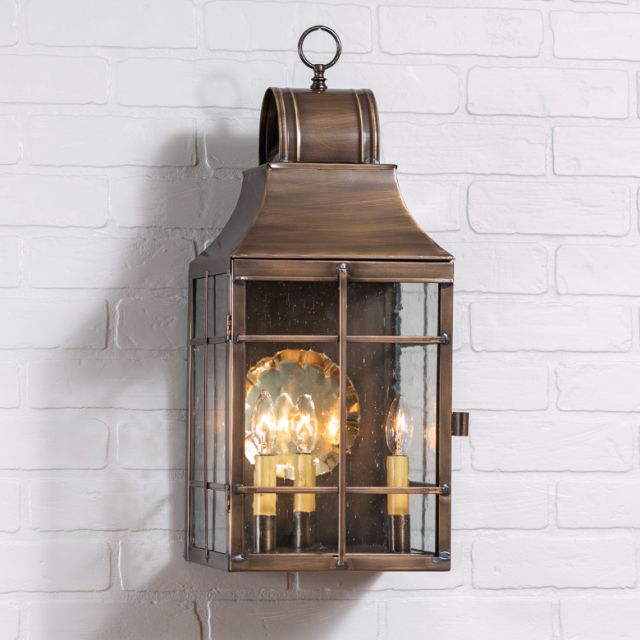 A solid weathered brass outdoor wall light with a blackened solder and lacquer coating, featuring a glass globe and three visible bulb sockets, mounted on a white brick wall.