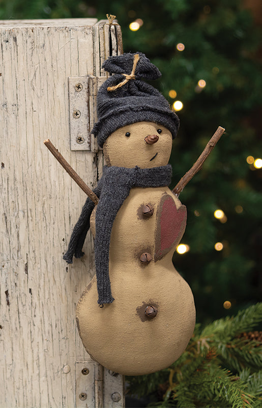 A decorative plush snowman with a carrot nose, jingle bell buttons, a red heart decal, and wearing a blue fabric hat and scarf, hanging on a wooden post with Christmas lights in the background.