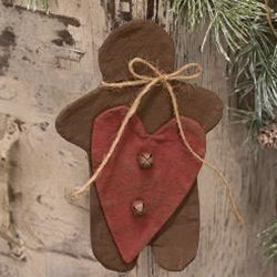 A stiffened brown fabric gingerbread man ornament with a large red heart decal and a jute bow. It is hanging against a backdrop of pine branches.