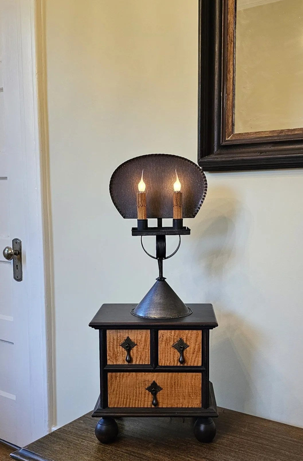 A rustic tin table light with a primitive finish, placed on a wooden table, with a warm glow emitting from the reflector.