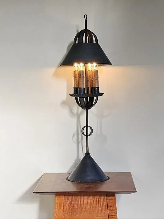A tall tin table light with a black finish and a geometric design, mounted on a wooden table against a plain wall.