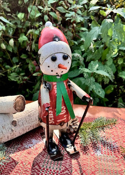 A decorative snowman figurine with a Santa hat, green scarf, and black boots, standing on a red and green surface with a log and greenery in the background.