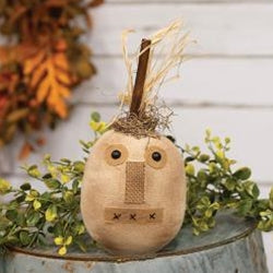 A stuffed toy resembling a pumpkin with a stitched face, moss detailing, and a top made of raffia, displayed on a blue surface with autumn leaves around it.