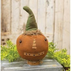 A stuffed toy resembling a pumpkin with a stitched patchwork face and a pointed green hat, set against a backdrop of greenery.