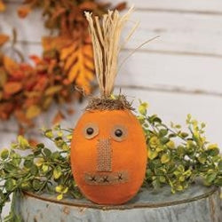 A decorative plush pumpkin with a stitched patchwork face and moss detailing, topped with raffia, placed on a blue surface with autumn leaves around it.