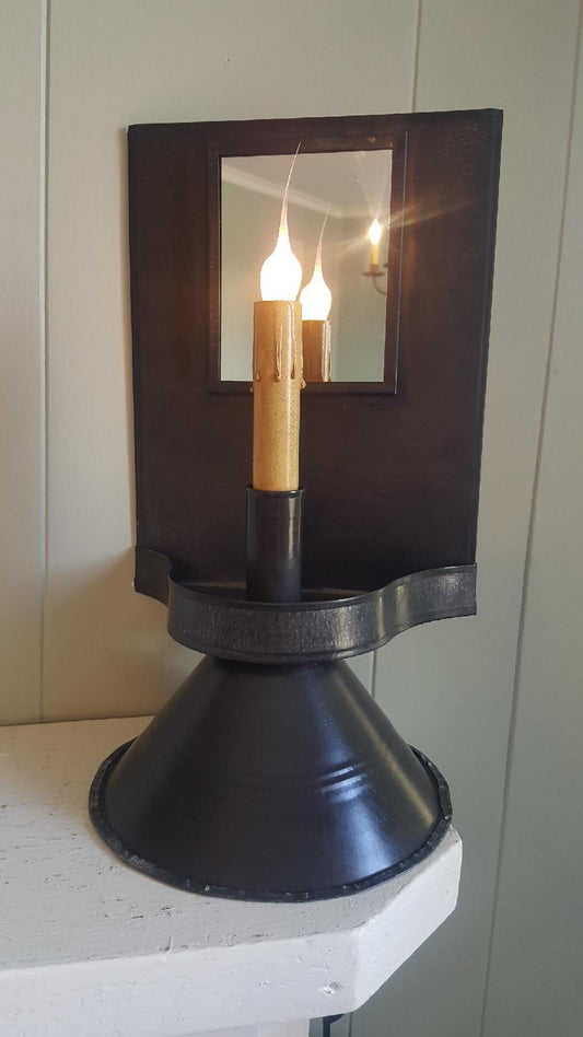 A tin table light with a warm glow, placed on a white surface with a mirror reflecting the light. The light has a rustic black finish and is mounted on a cylindrical base.