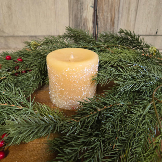 A pillar candle with a yellow flame, surrounded by green pine branches and red berries, hand-poured and placed on a wooden surface.