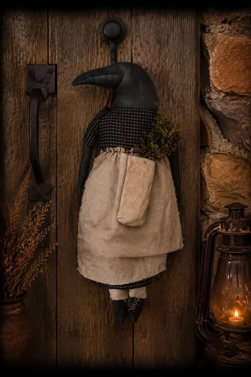 A rustic black raven doll with a beige apron and black-and-white check shirt hangs on a dark wooden door, with a stone wall, lantern, and dried flowers in the background. Text at the bottom reads 'Primitive Kreations Wholesale'.