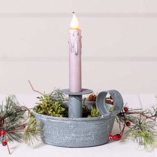 A weathered zinc candle holder with a loop-style handle, featuring a two-tiered design. The lower pan is filled with greenery, and a candle is placed on the upper pan.