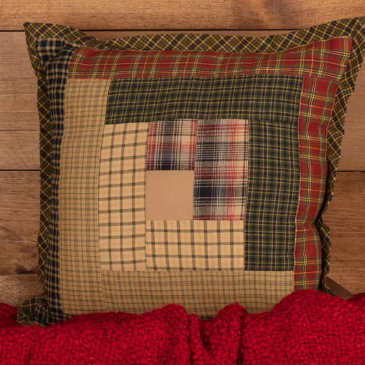 A square patchwork throw pillow with a rustic plaid and checkered design, styled on a wooden surface with a red knit blanket underneath.