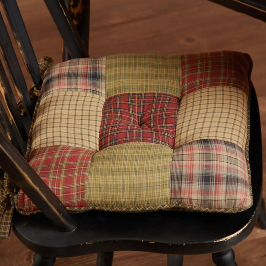 A square patchwork tufted cushion with a rustic plaid pattern, shown resting on a dark wooden chair over a wood floor.