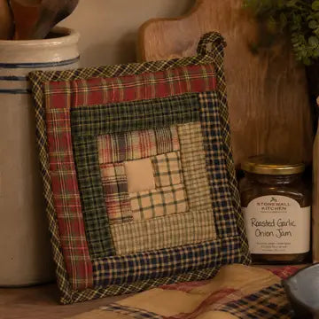 A square quilted fabric pot holder with a rustic patchwork plaid design in red, green, beige, and blue, styled on a kitchen counter next to a jar of roasted garlic onion jam.