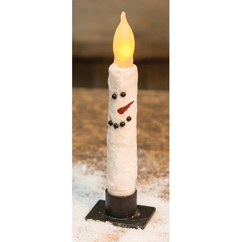 A white taper candle with a snowman face, lit with a warm glow, placed on a black base with snowflakes around.