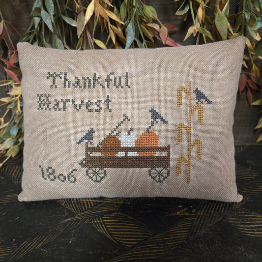 Decorative pillow with cross-stitched design of a harvest scene and text, placed on a textured surface with greenery.