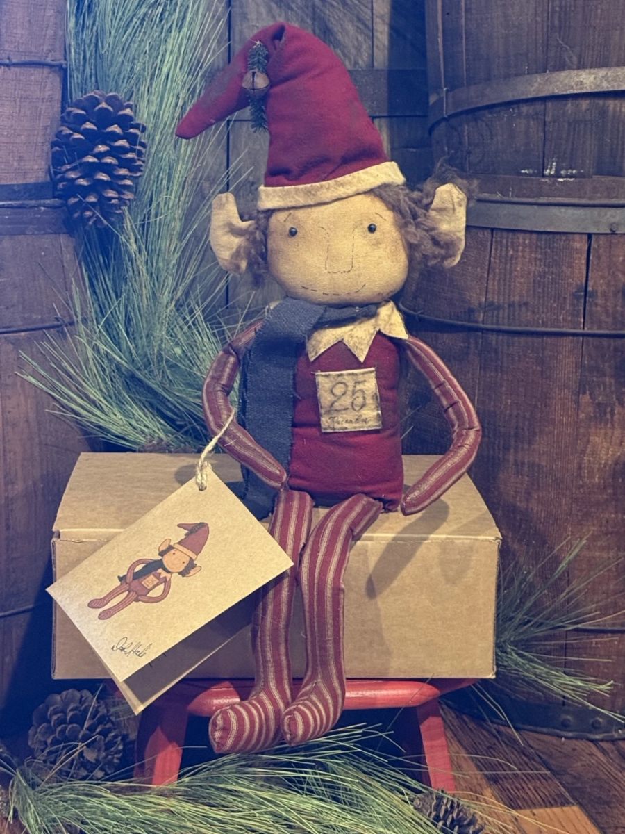 A rustic Christmas elf doll with a red hat, red striped legs, and a dark blue scarf, sitting on a cardboard box with a hanging gift tag in a holiday-themed setting.