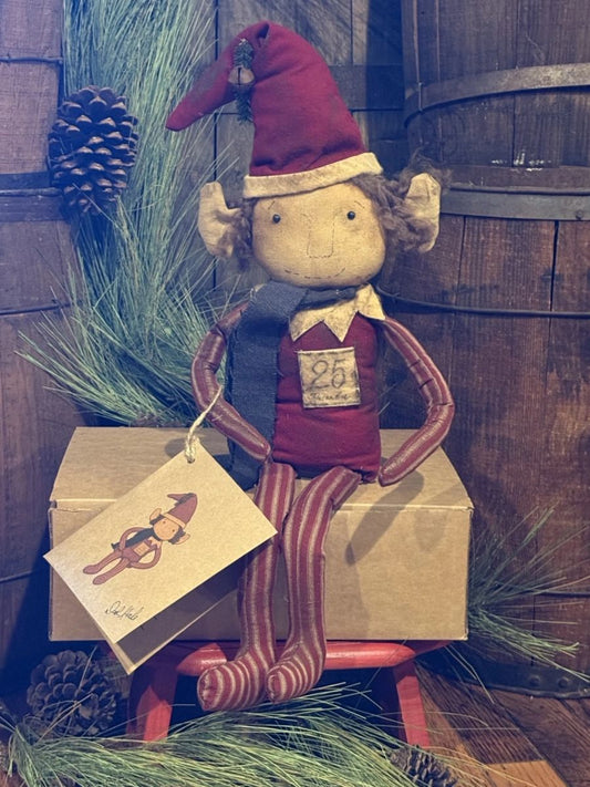A rustic Christmas elf doll with a red hat, red striped legs, and a dark blue scarf, sitting on a cardboard box with a hanging gift tag in a holiday-themed setting.