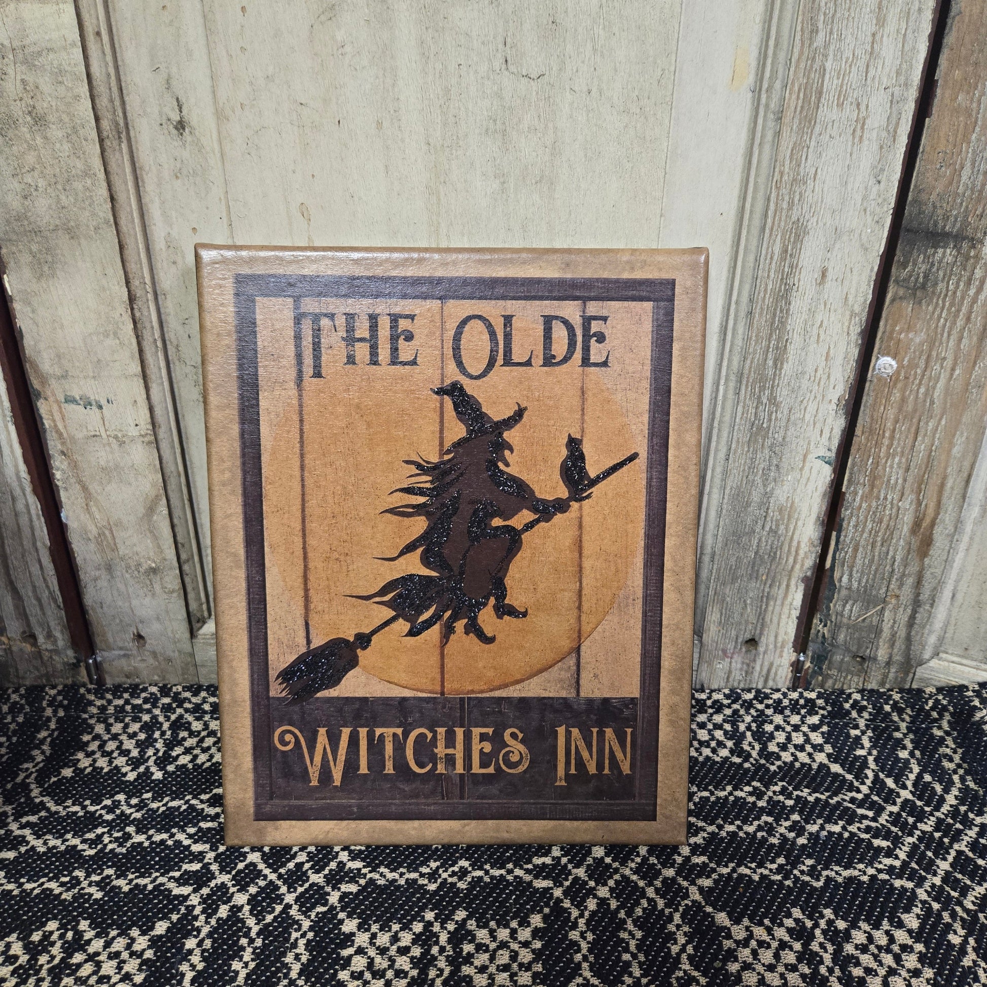 A canvas print titled 'The Olde Witches Inn' featuring an illustration of a witch on a broomstick with a crescent moon in the background, framed in a wooden border.