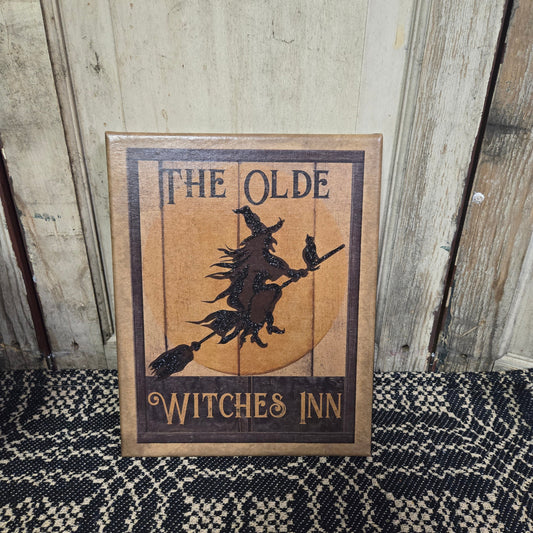 A canvas print titled 'The Olde Witches Inn' featuring an illustration of a witch on a broomstick with a crescent moon in the background, framed in a wooden border.