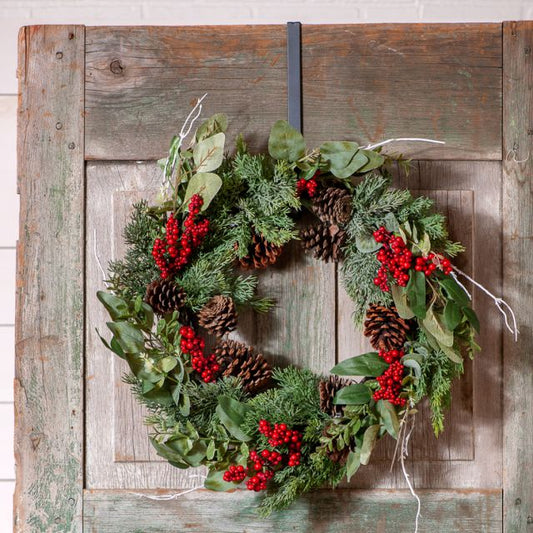 A wreath hanging on a wooden door, featuring a combination of greenery, red berries, and pine cones.