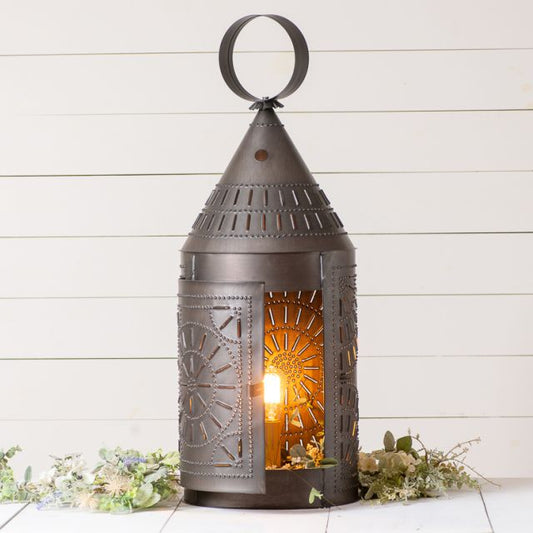 A black, iron lantern with a geometric pattern, featuring a chisel in the top and a lit candle inside, placed on a white surface with decorative elements around it.
