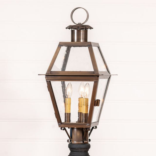 An outdoor post light with a solid weathered brass base and a glass cover, featuring three light bulbs inside, mounted on a circular black post.
