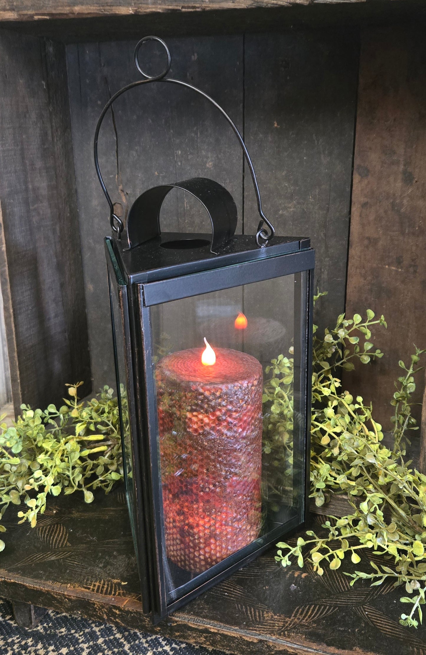 Black lantern with a lit candle inside, surrounded by greenery on a wooden surface.